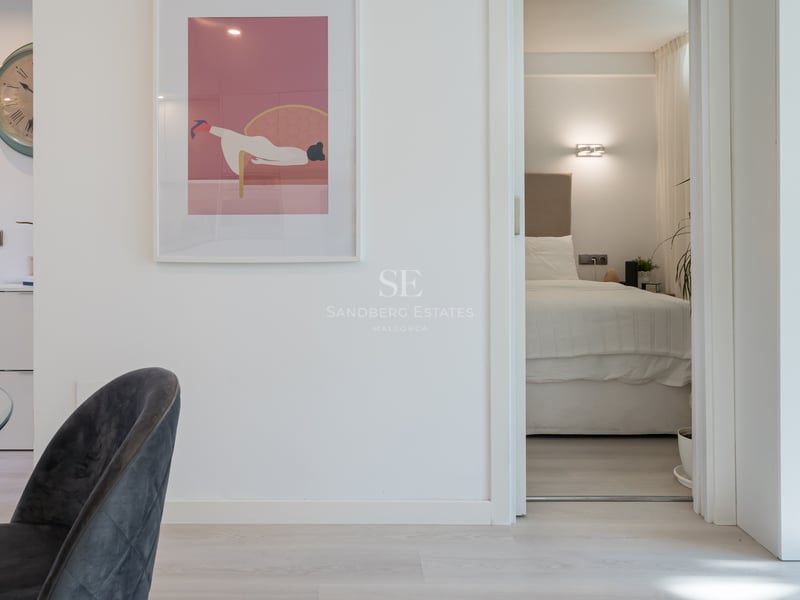 View through an open white door into a minimalist bedroom with a large bed, beige headboard, and warm wall lighting.