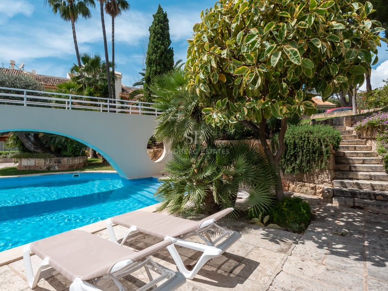Sun loungers by a turquoise pool with a white arch bridge and lush landscaping.