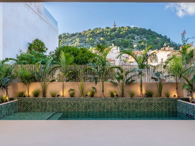 Piscine moderne avec carreaux verts en chevrons entourée de plantes tropicales et vue sur une colline sous un ciel bleu.