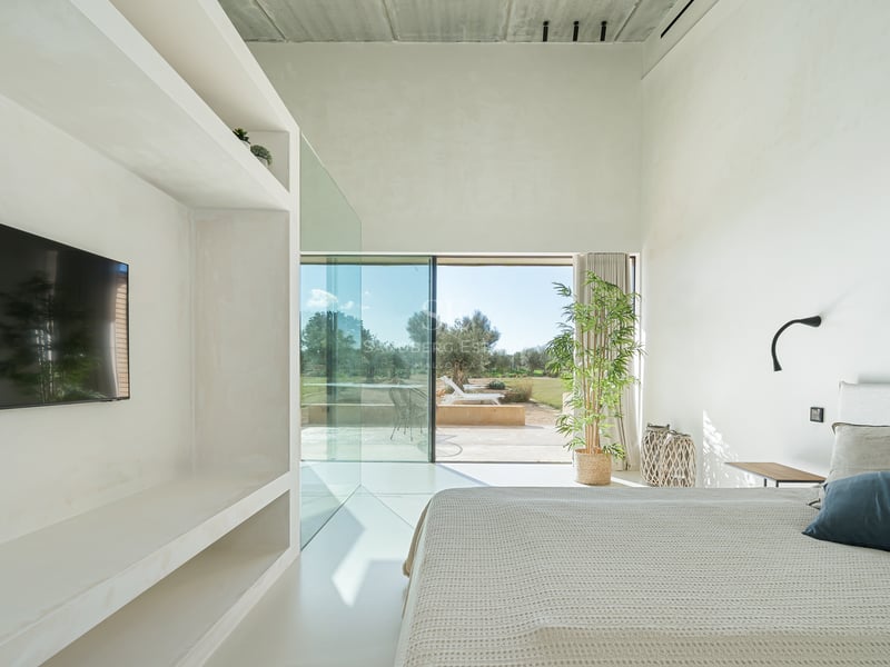 Minimalistiskt vitt sovrum med stora glaspartier som vetter mot en solig terrass och trädgård.