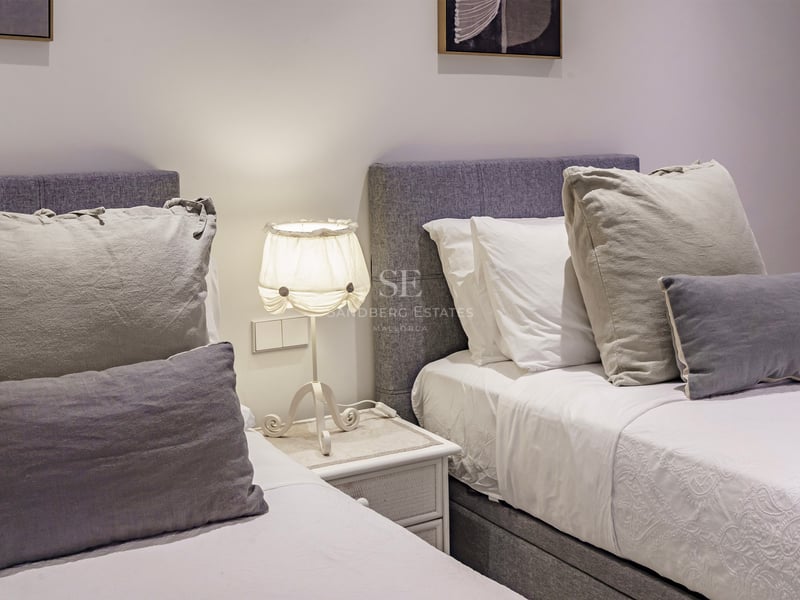 Two twin beds with grey upholstered headboards, white linens, and a lamp on a bedside table between them.