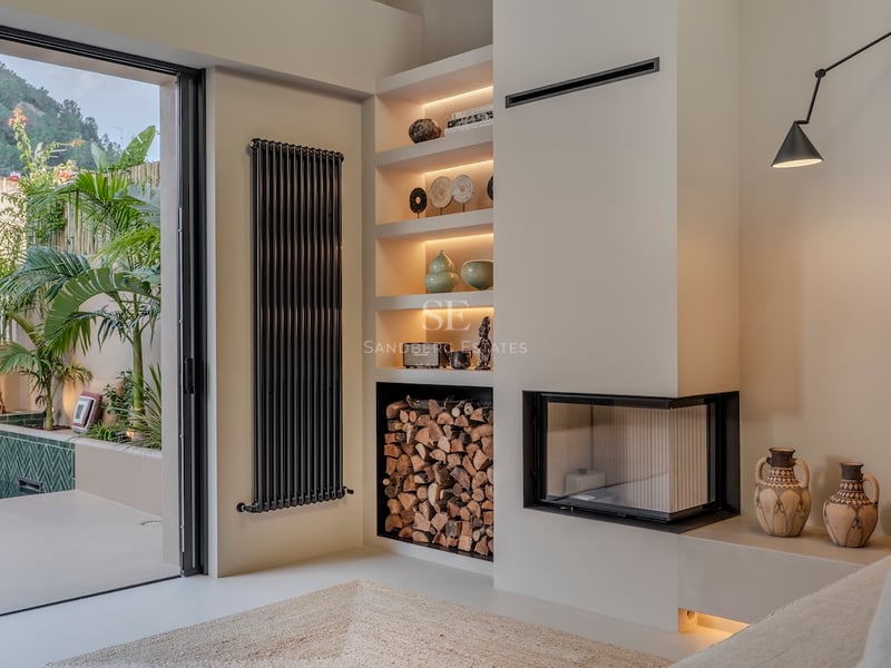 Espace de vie intérieur avec cheminée d'angle moderne, rangement à bois et vue sur une terrasse extérieure luxuriante.