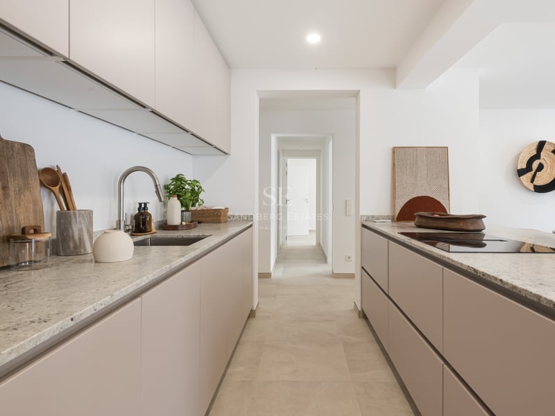 Cuisine moderne avec des placards beiges, des plans de travail en pierre et une vue sur un couloir blanc.