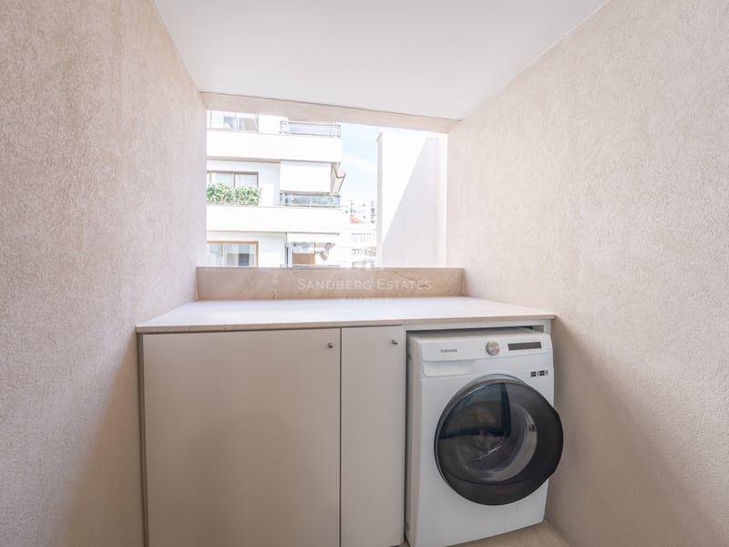 En modern tvättmaskin och vita skåp inbyggda i en beige balkongnisch med utsikt över stadsbyggnader.