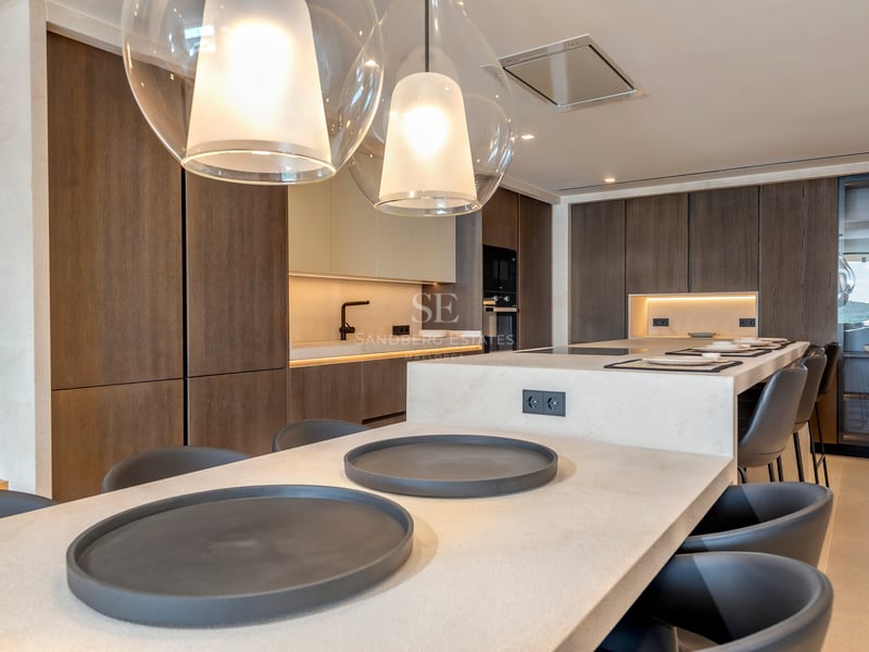 Cuisine moderne avec placards en bois sombre, îlot en pierre claire avec taburetes et suspensions en verre.