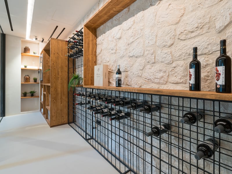 Modern vinkällare med vit stenmur, svarta metallställ för flaskor och detaljer i ljust trä.