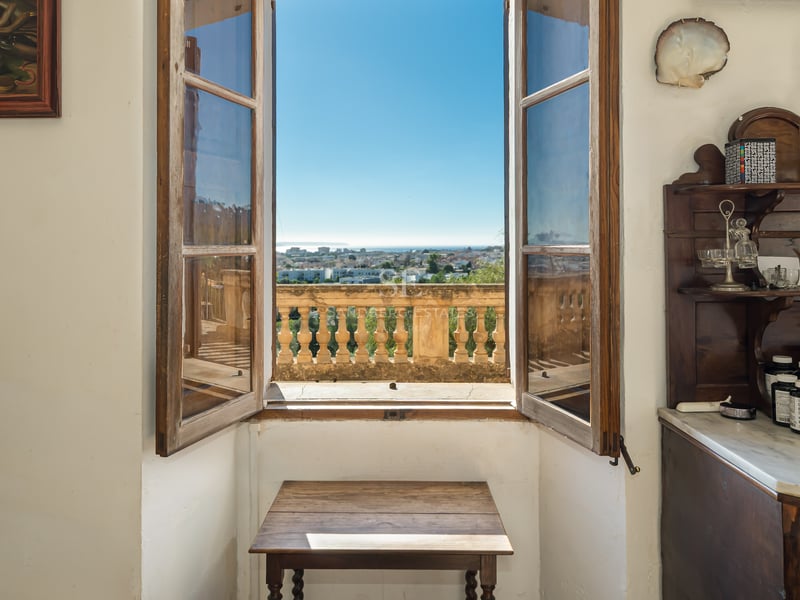 Fenêtre en bois ouverte donnant sur une ville côtière et la mer sous un ciel bleu clair.