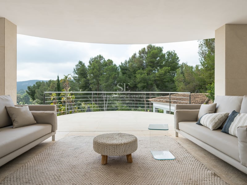 Terrasse moderne avec canapés gris et pouf tressé donnant sur une forêt dense de pins.