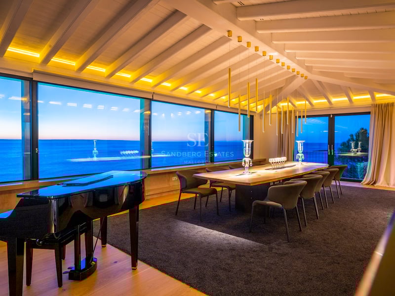 Ein modernes Esszimmer mit Flügel, großem Holztisch und bodentiefen Fenstern mit Blick auf das Meer in der Dämmerung.