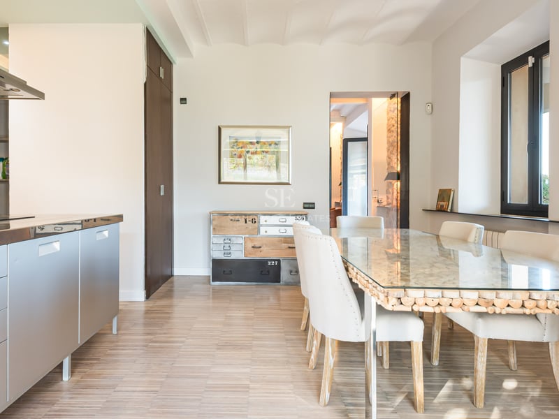 Ljus matsal med glasbord, vita stolar och kök i rostfritt stål.