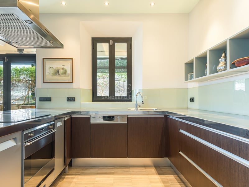 Modernt kök med mörka träskåp, bänkskivor i rostfritt stål och stänkskydd i glas under ljus belysning.