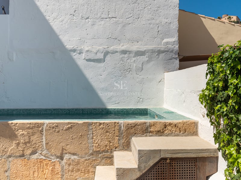 Petite piscine surélevée avec mosaïque turquoise et socle en pierre naturelle contre un mur blanc et du lierre.