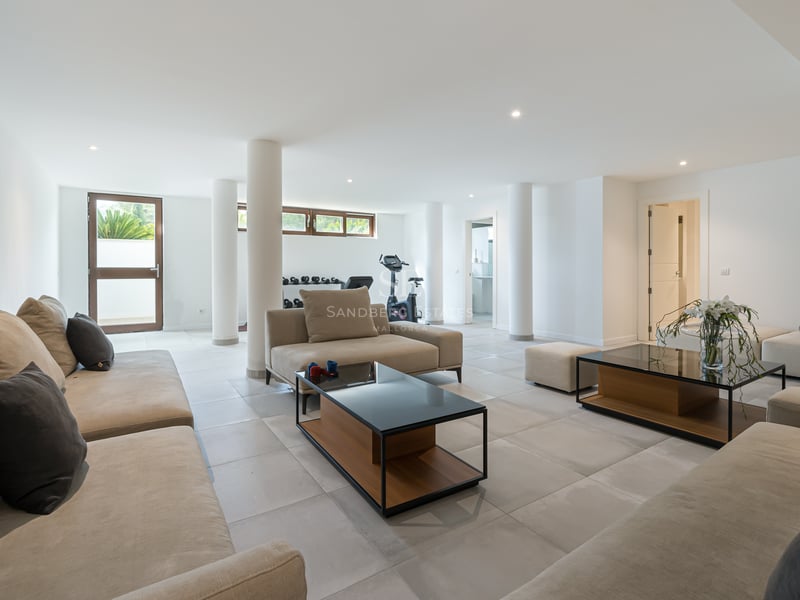 Grande pièce lumineuse avec canapés taupe, sol en carrelage et coin fitness avec vélo d'appartement.