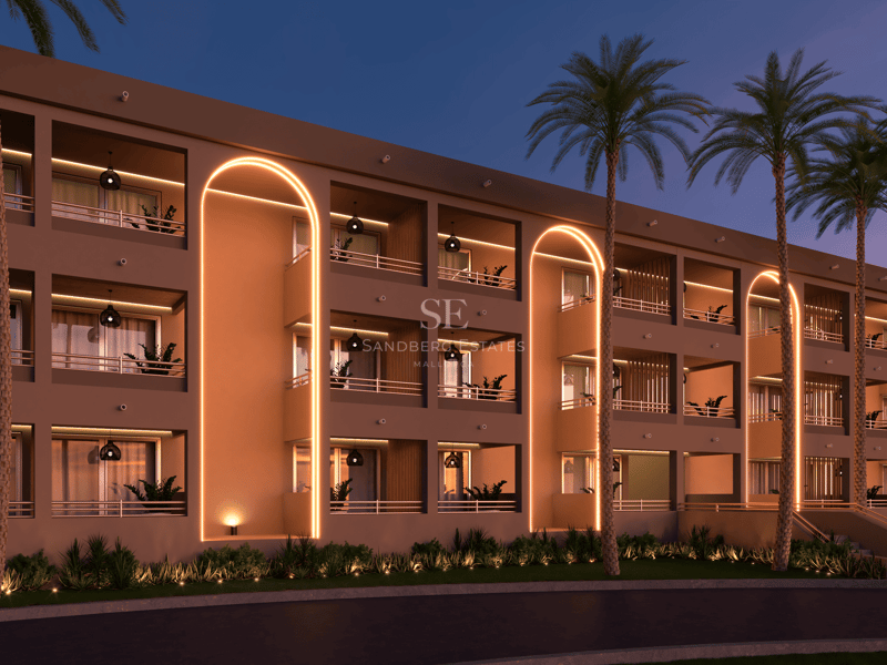 Bâtiment moderne avec arches dorées illuminées, balcons privés et palmiers sous un ciel de crépuscule.