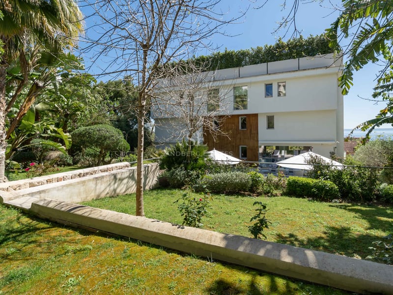 Moderne weiße Fassade mit Holzverkleidung, Blick auf einen grünen Garten und Poolbereich mit Sonnenschirmen.