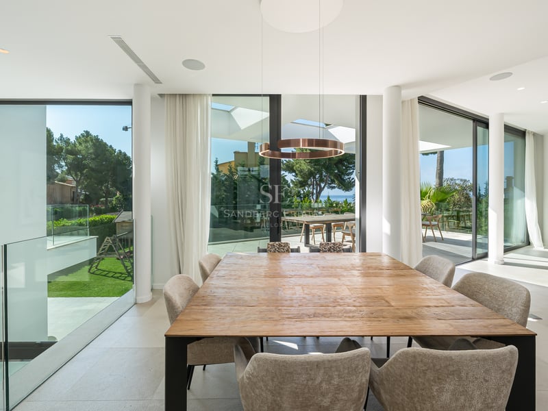 Grande table à manger en bois avec chaises beiges dans une pièce lumineuse avec baies vitrées et vue sur la mer.