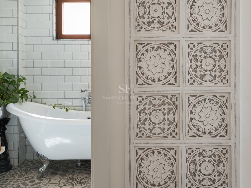 Salle de bain avec baignoire à pattes blanche, carrelage métro et sol à motifs, vue à travers une porte en bois sculpté.