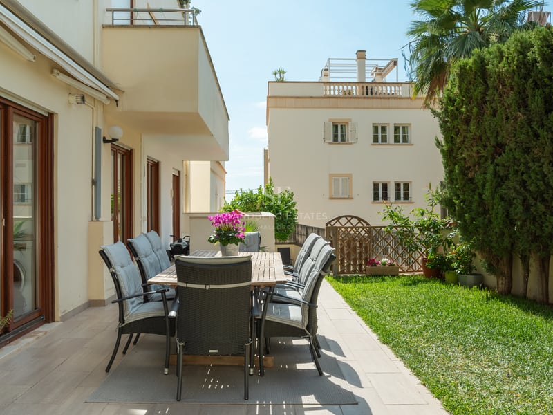 Espace repas extérieur sur terrasse carrelée bordant un jardin avec pelouse et haies hautes sous le soleil.
