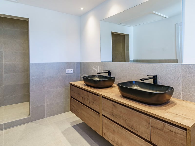 Samtida badrum med grått kakel, dubbelkommod i trä, svarta tvättställ och en rymlig duschkabin.