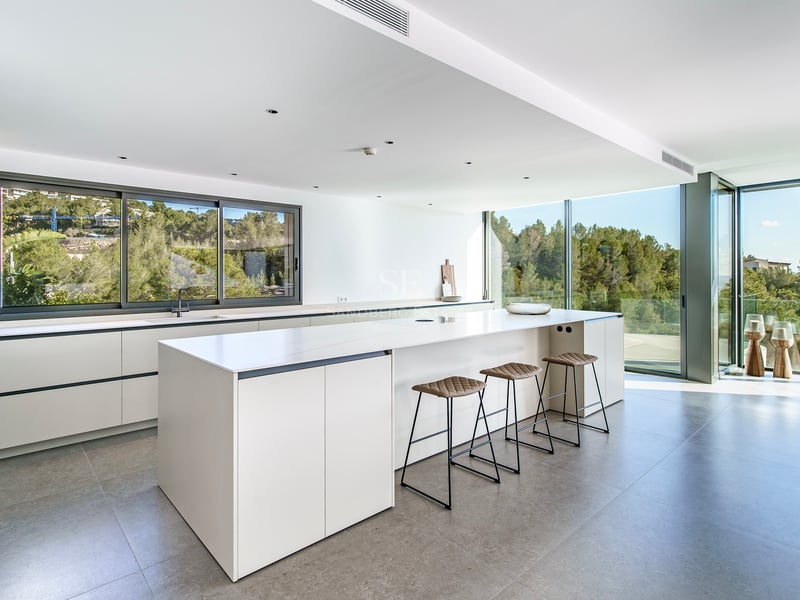 Cuisine lumineuse avec îlot blanc, tabourets en cuir et grandes baies vitrées donnant sur une forêt.