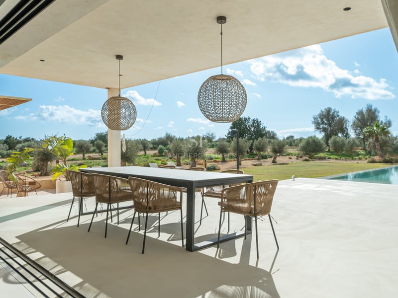 Övertäckt matplats utomhus med modernt bord och flätade stolar med utsikt över en trädgård med olivträd.