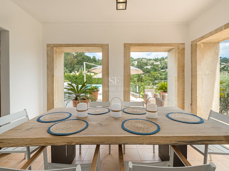 Table à manger en bois face à des ouvertures en pierre offrant une vue sur un jardin verdoyant et une piscine.