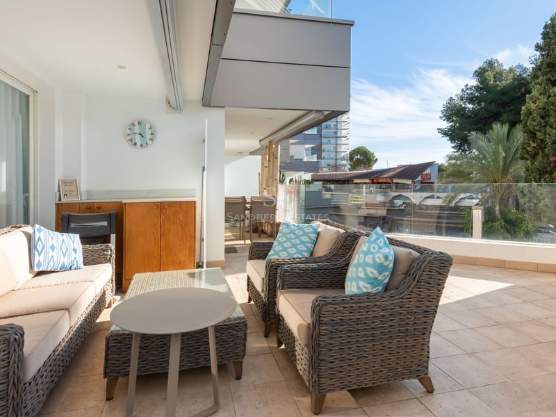 Grande terrasse avec canapés en osier gris, coussins bleus et garde-corps en verre sous un ciel bleu.