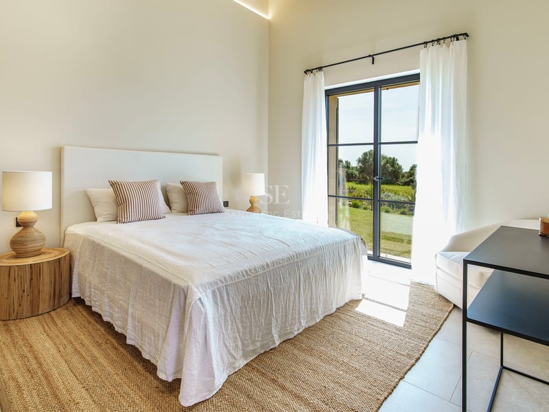 Helles Schlafzimmer mit weißem Bett, Jute-Teppich und bodentiefen Fenstern, die ins Grüne führen.