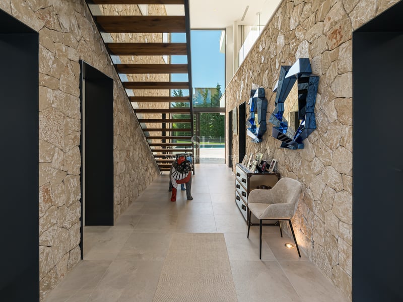 Couloir moderne avec murs en pierre, escalier flottant en bois et grandes baies vitrées vue sur le jardin.