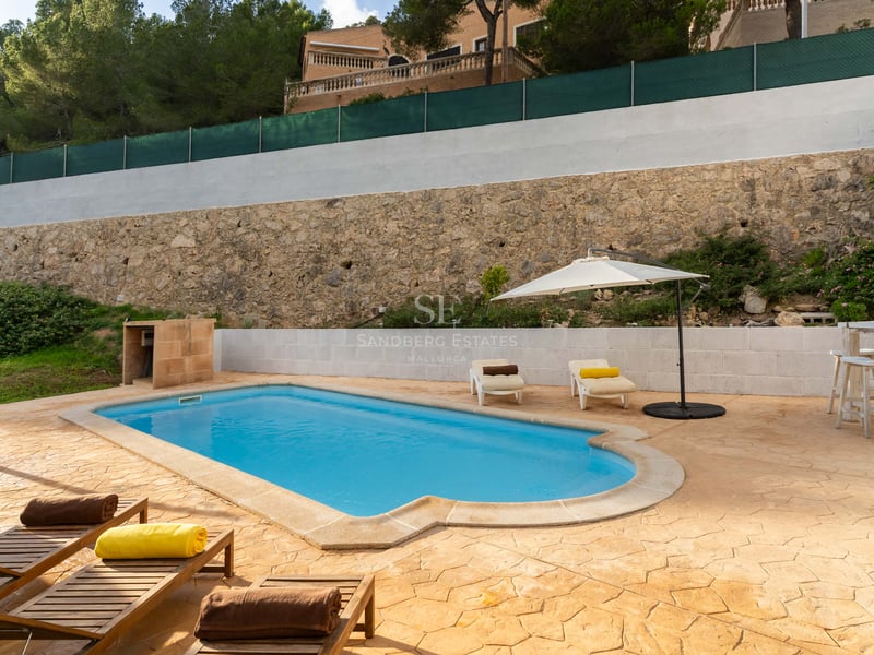 Piscine extérieure avec terrasse en pierre, chaises longues, parasol blanc et mur de pierre.