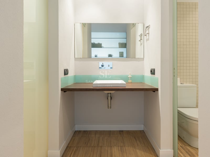 Badrum med svävande bänkskiva i trä, vitt fyrkantigt handfat, spegel och turkos glaspanel.