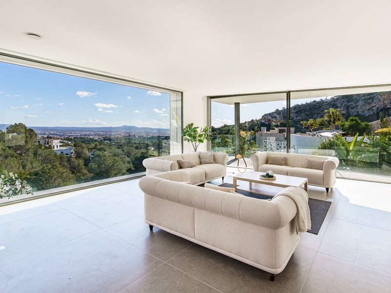 Salon moderne avec trois canapés beiges et de grandes baies vitrées offrant une vue sur la ville et les montagnes.