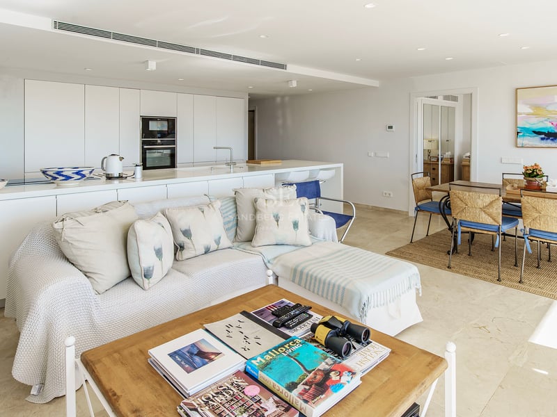 Bright open-plan living room with white sofa, wooden coffee table, dining set, and minimalist kitchen in the background.