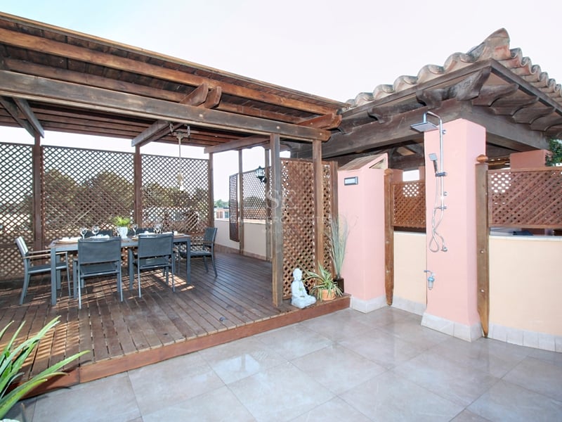 En träpergola över en matgrupp på ett mörkt trädäck, med en utedusch monterad på en rosa vägg.