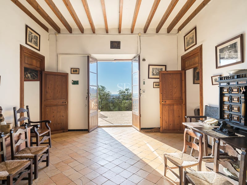 Hall traditionnel avec poutres en bois, sol en terre cuite et portes ouvertes donnant sur une terrasse ensoleillée.