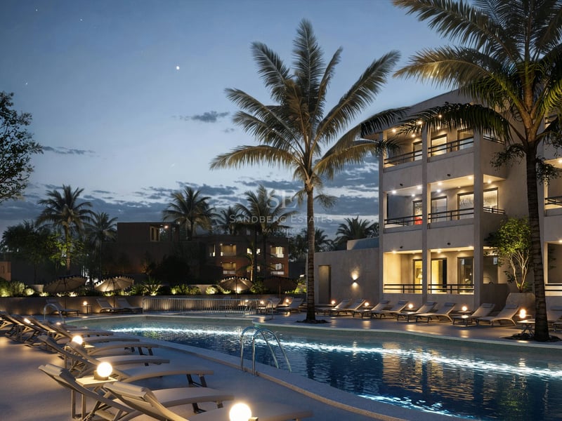 Piscine illuminée la nuit avec des palmiers, des chaises longues et un bâtiment moderne à plusieurs étages.