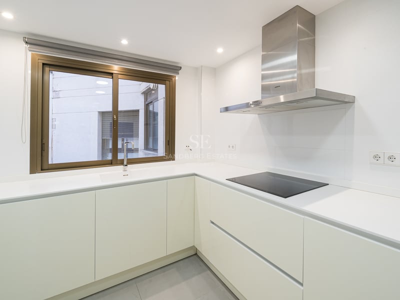 Minimalistiskt vitt kök med L-formad bänkskiva, induktionshäll och köksfläkt i rostfritt stål.