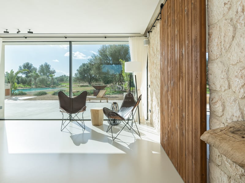 Ljust vardagsrum med stora glaspartier, butterfly-stolar, skjutdörr i trä och vägg i natursten.