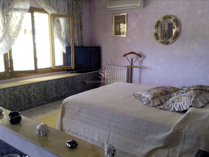 Dormitorio con paredes de color lila, cama grande con cojines florales y amplios ventanales de madera bajo luz natural.