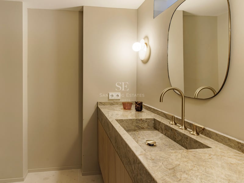 Nahaufnahme eines Waschbeckens aus Naturstein mit goldenem Wasserhahn, ovalem Spiegel und warmer Wandbeleuchtung.