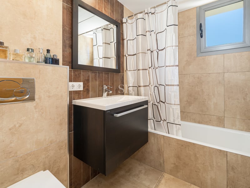 Modernt badrum med beige kakel, mörk kommod, badkar med draperi och ett litet fönster.