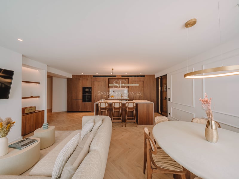 Grand salon luxueux avec canapé beige, parquet en chevrons et cuisine intégrée en bois naturel.