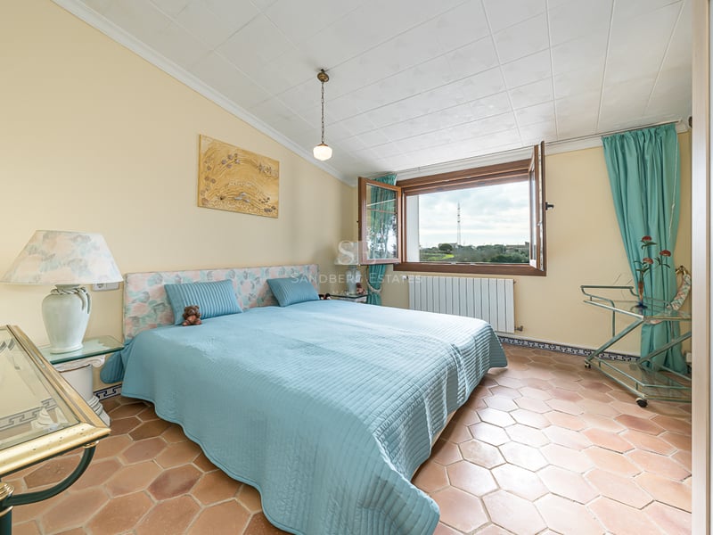 Chambre avec sol en terre cuite, lit bleu et fenêtre ouverte donnant sur un paysage verdoyant.