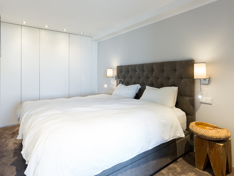 Zeitgenössisches Schlafzimmer mit weißem Bett, grauem Polsterkopfteil, weißen Wandschränken und Designer-Holzhocker.