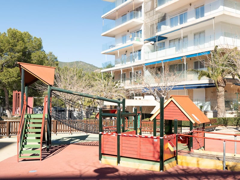 Kinderspielplatz mit Holzgeräten und Sicherheitsboden vor einem modernen Apartmenthaus.