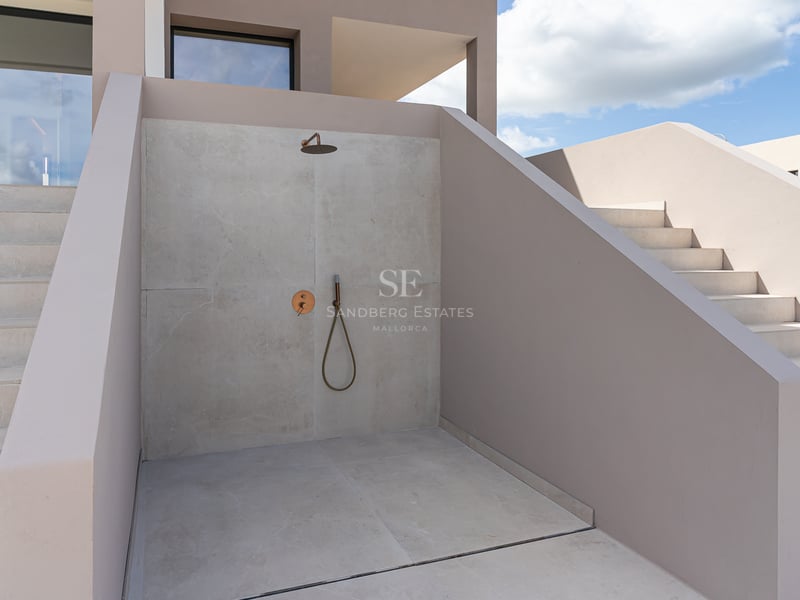 Minimalistische Außendusche mit hellgrauen Mikrozementwänden und Messingarmaturen vor blauem Himmel.