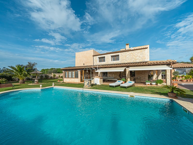 Grande piscine turquoise devant une villa méditerranéenne traditionnelle en pierre avec pelouse et palmiers.