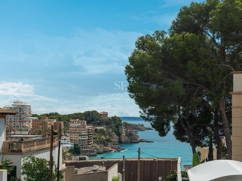 Vue surélevée d'une baie turquoise avec des bâtiments méditerranéens, des falaises rocheuses et un grand pin.