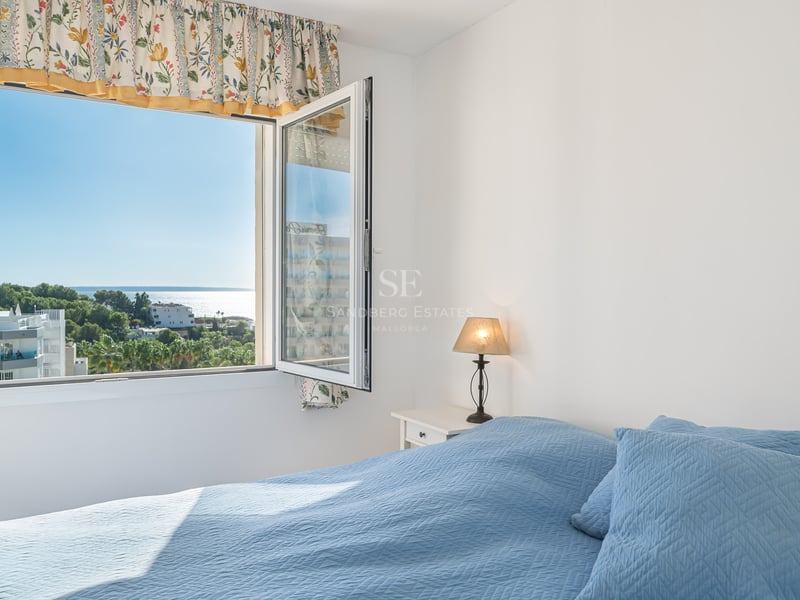 A sunlit bedroom featuring a blue quilted bed and an open window looking out over trees and the sparkling sea.