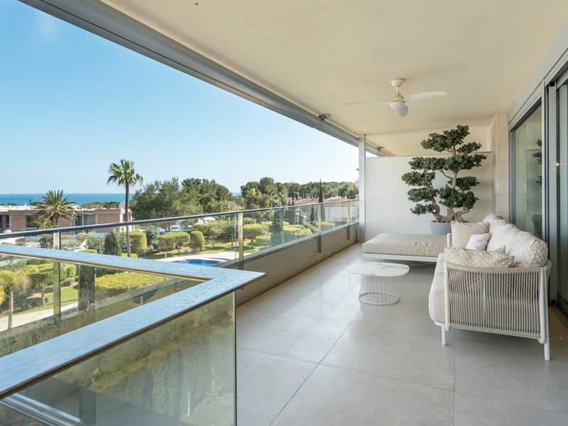 Wide modern terrace with white outdoor sofas, glass railings, and views of lush gardens and the sea in the distance.